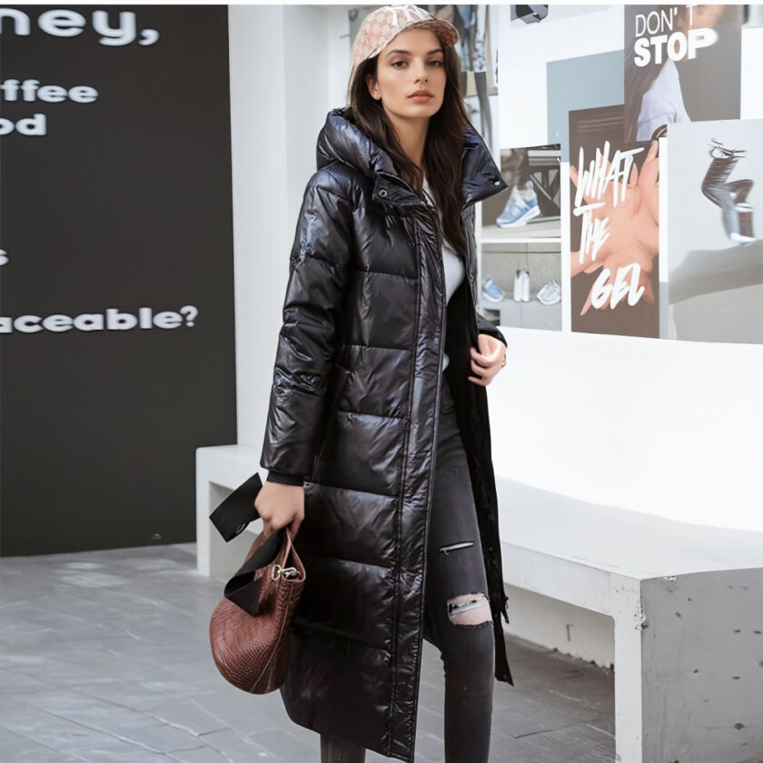 Femme debout avec en arrière plan, un mur avec des inscriptions sur la droite et un mur noir à gauche avec des inscriptions blanches. Un banc en pierre grise derrière la femme. Elle porte un pantalon dlim noir troué au genou gauche, une casquette beige, une doudou longue noire à capuche et un sac à main dans sa main droite.