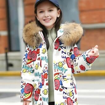 Jeune fille dans une rue portant une doudoune à capuche blanche pour ado La doudoune a une capuche en fourrure beige Des motifs colorés sont imprimés sur l'ensemble du manteau La jeune fille porte une casquette bleu marine.