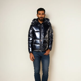 Homme brun debout contre un mur beige, portant un jean bleu denim et une doudoune brillante à capuche de couleur noire.