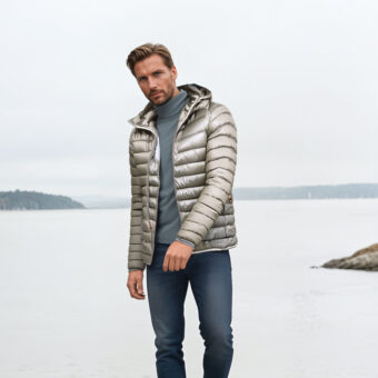 Homme debout dos à un plan d'eau portant un jean bleu, un haut gris et une doudoune courte fine et légère avec capuche beige.