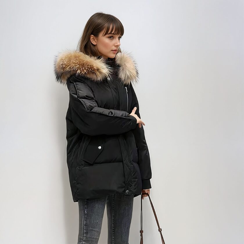 Femme brune avec une frange, devant un mur blanc/gris, portant une doudoune noire à capuche avec fourrure marron mi-longue matelassée. Elle tient la sangle d'un sac à main dans sa main gauche.