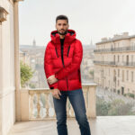 Homme brun debout sur une terrasse en ville portant un jean bleu et une doudoune matelassée rouge avec capuche. Au fond on voit un bâtiment style immeuble parisien.
