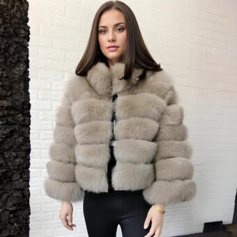 Femme aux cheveux châtains debout dos à un mur en pierres blanches, portant une doudoune en fausse fourrure beige et un pantalon noir.