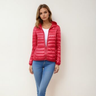 Femme blonde debout dos à un mur beige, portant un jean slim bleu, un haut blanc et par-dessus, une doudoune légère et chaude rouge, fermée avec sa fermeture éclair à un tiers laissant apparaître le haut blanc.