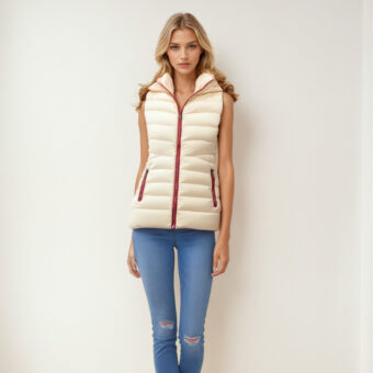 Femme blonde aux cheveux détachés portant un jean bleu et une doudoune légère sans manches blanche avec les coutures rouges. Elle est debout dos à un mur beige.