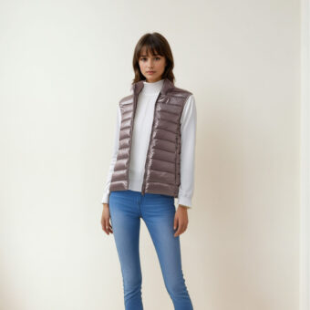 Femme brune avec frange portant un haut à manches longues blanc avec une doudoune légère et chaude sans manches beige et un pantalon jean bleu. Debout dos à un mur beige.