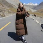 Femme portant une doudoune longue marron. Elle est debout sur une route au milieu des montagnes.