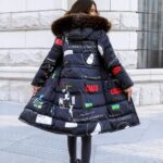 Femme brune debout de dos avec un mur en pierres beiges face à elle, la tête tournée vers la droite. Elle porte un pantalon noir et une doudoune réversible et longue de couleur bleu marine avec des motifs et des inscriptions de couleur blanche, rouge et verte. La capuche a de la fourrure sur le bord.