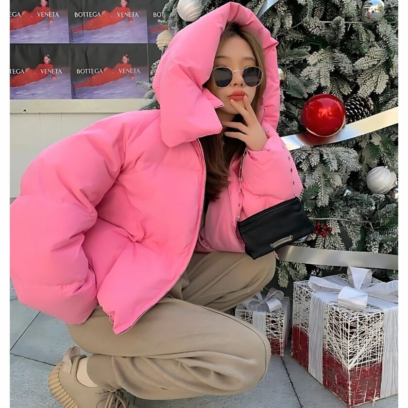 Femme accroupie avec lunettes de soleil devant un sapin de Noël avec une doudoune rose, capuche sur la tête.