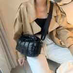 Femme assise avec un pantalon blanc, un haut noir et une veste en cuir camel, avec un sac noir brillant matelassé en bandoulière