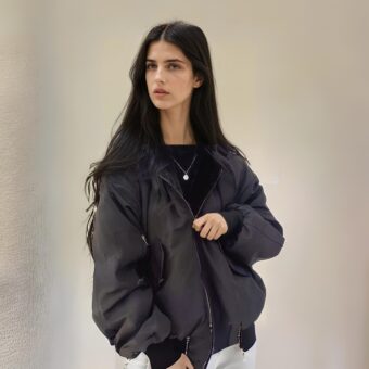 Jeune femme mince et brune, aux cheveux longs détachés, portant une veste réversible noire. Debout dos à un mur beige.