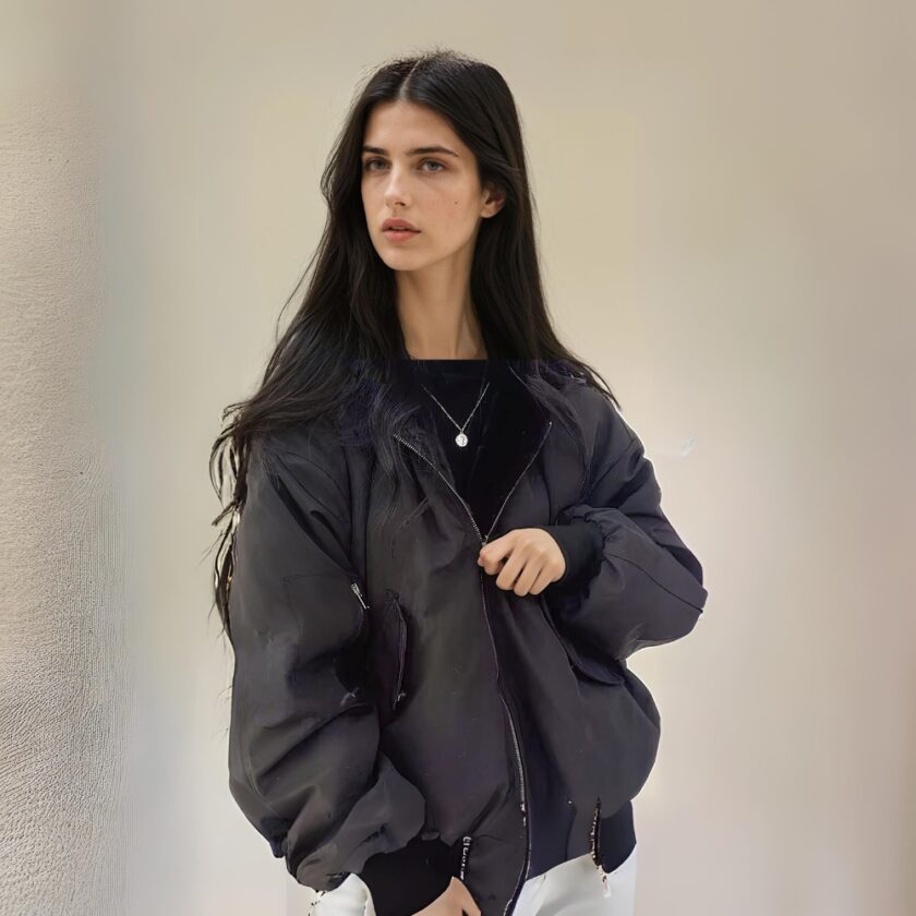 Jeune femme mince et brune, aux cheveux longs détachés, portant une veste réversible noire. Debout dos à un mur beige.