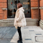On voit une jeune femme brune qui est debout dans la rue. Elle se tient de côté et porte un jean noir avec des chaussures lacées montantes assorties et une doudoune de grossesse beige ajustable grâce à des cordons.