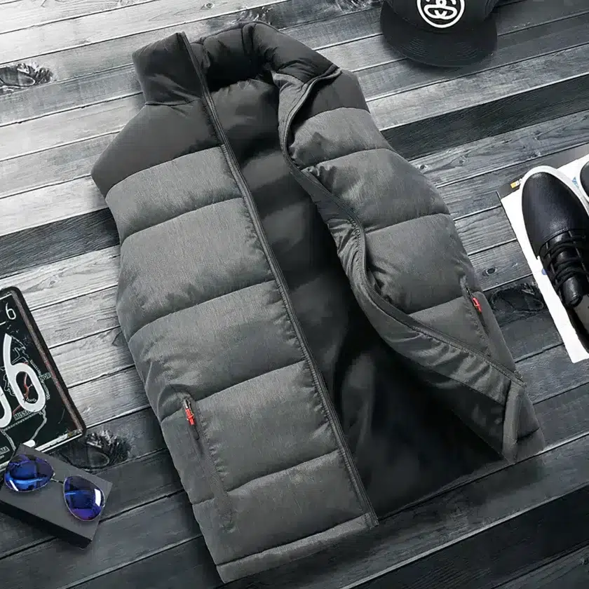 Doudoune grise et noir sur le haut du buste matelassée sans manches avec poches zippées semi-ouverte étendue sur du parquet à latte gris foncé. Sur les côtés , il y a une casquette noire, une basket noire et un bout de plaque d'immatriculation.