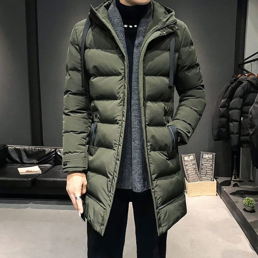 Homme portant une doudoune longue et matelassée verte. Fond noir et parquet clair.