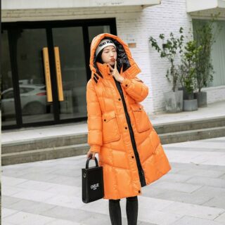 Femme portant une doudoune orange et un sac à main noir dans la main, se tenant dans la rue.