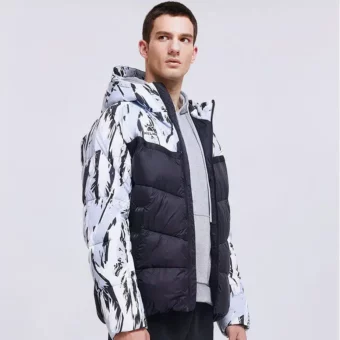 Homme brun debout sur fond gris portant une doudoune de foot courte ouverte laissant apparaitre un sweat gris. La doudoune est en noir matelassé uni sur le bas du buste et tigré en noir et blanc sur le haut des épaules et les manches.