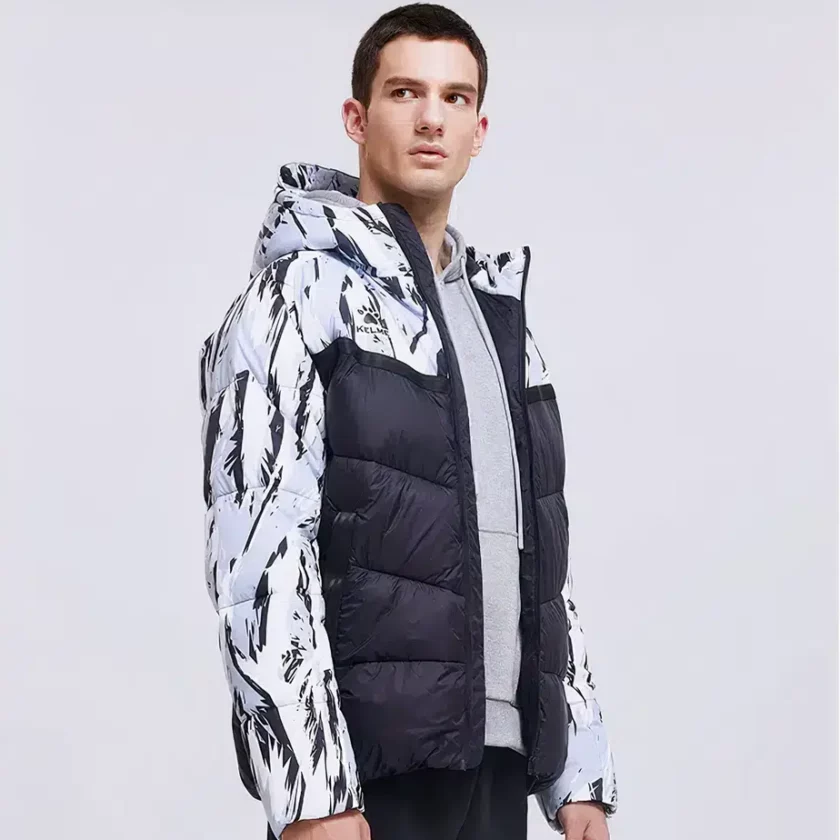 Homme brun debout sur fond gris portant une doudoune de foot courte ouverte laissant apparaitre un sweat gris. La doudoune est en noir matelassé uni sur le bas du buste et tigré en noir et blanc sur le haut des épaules et les manches.