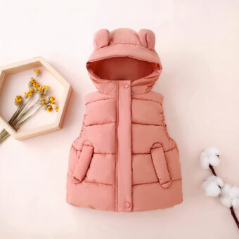 On voit une doudoune rose sans manche avec capuche à oreilles pour petite fille. Elle est présentée sur un fond rose pastel près de fleurs de coton et d'un bouquet de fleurs jaunes dans un cadre