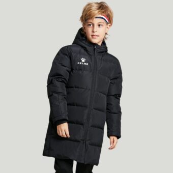 Un jeune garçon blond porte la doudoune longue de sport en coton rembourré pour enfant, et il pose devant un fond gris.