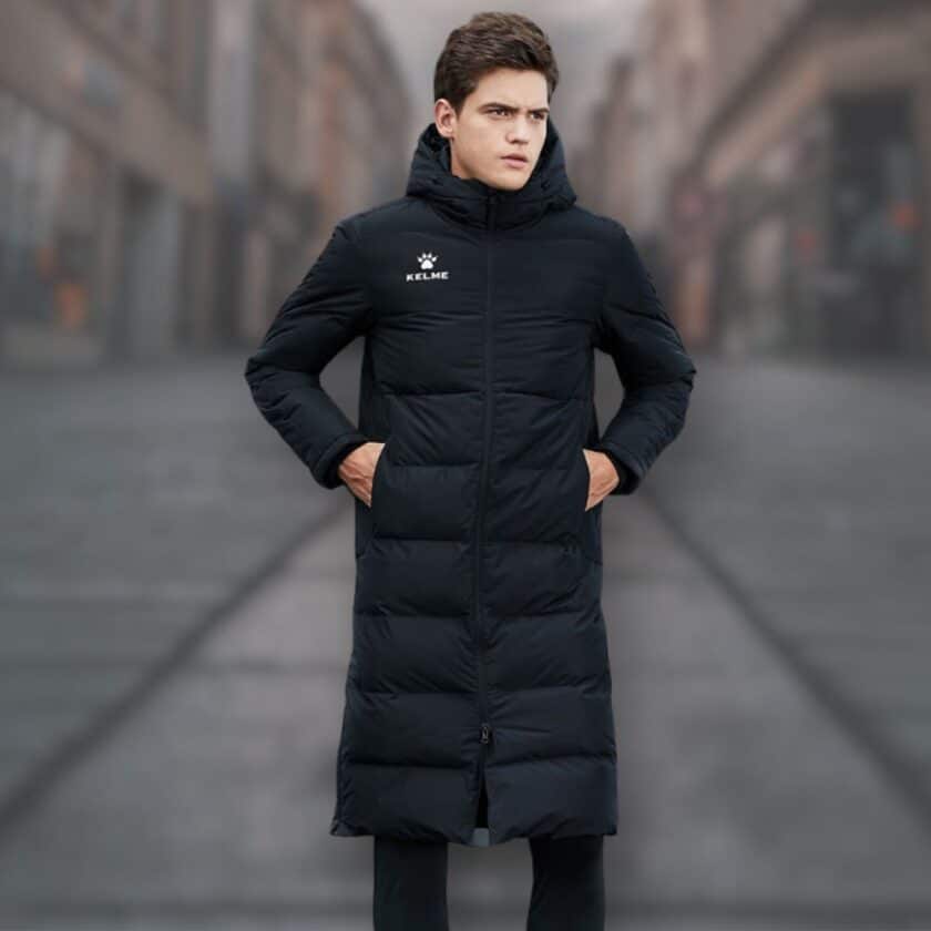 Doudoune de foot noire unisexe longue et imperméable portée par un jeune homme brun. Il semble se trouver dans une rue.