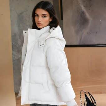 Femme brune de profil portant une doudoune courte à capuche en duvet de couleur blanche. Un sac à main derrière elle et un mur beige et gris.