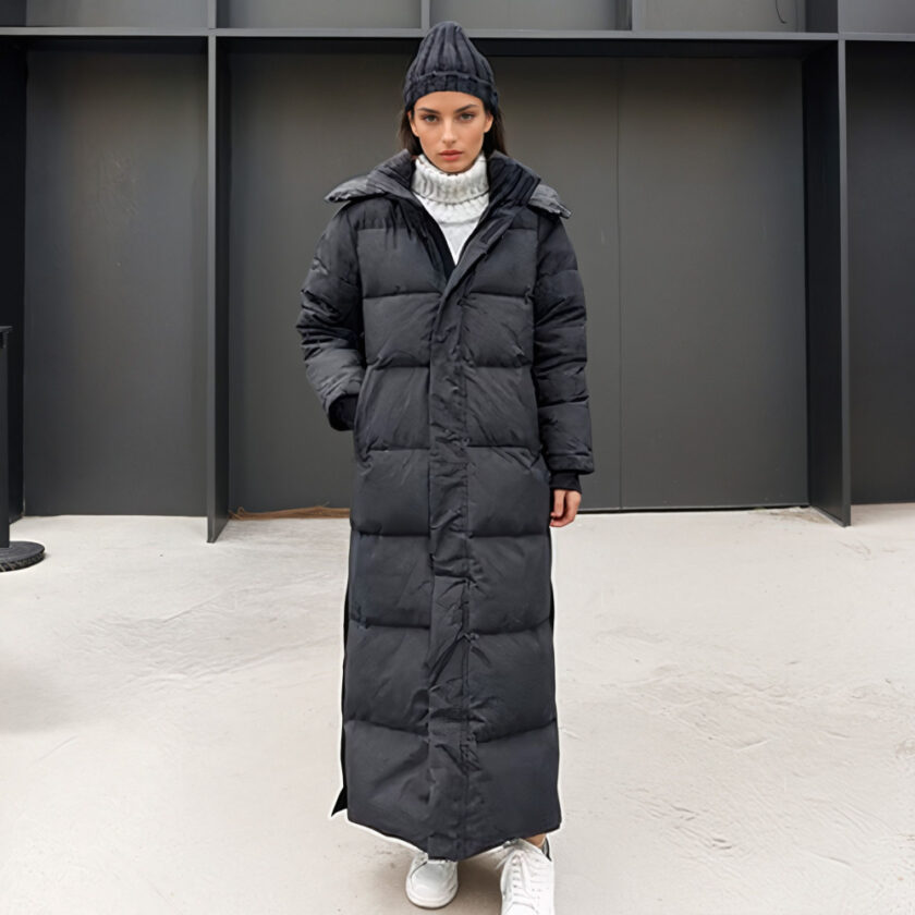 Femme brune debout avec des chaussures blanches, un bonnet en laine noir et une doudoune extra longue matelassée noire. Sa main droite dans la poche de doudoune. Au fond, un mur noir style en bois.