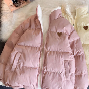On voit deux doudounes : une rose clair et une beige. Elles arborent toutes les deux un petit cœur brodé sur la poitrine. Elles sont réversibles et l'autre face est en polaire avec un motif ourson sur la poitrine. Elles sont présentés sur un tapis en faux mouton, près d'un casque audio. La photo est prise de haut.