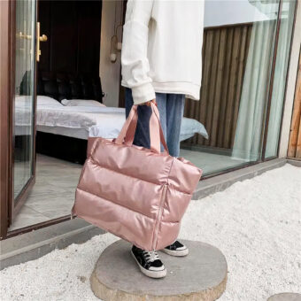 Grand sac doudoune matelassé rose métallisé porté par une personne à la main. La personne vêtue d'un sweat blanc et d'un jean bleu est debout à l'extérieur sur un fin rondin de bois sur un sol de gravier blanc. On distingue un décor de chambre avec un lit en arrière-plan derrière une baie vitrée.