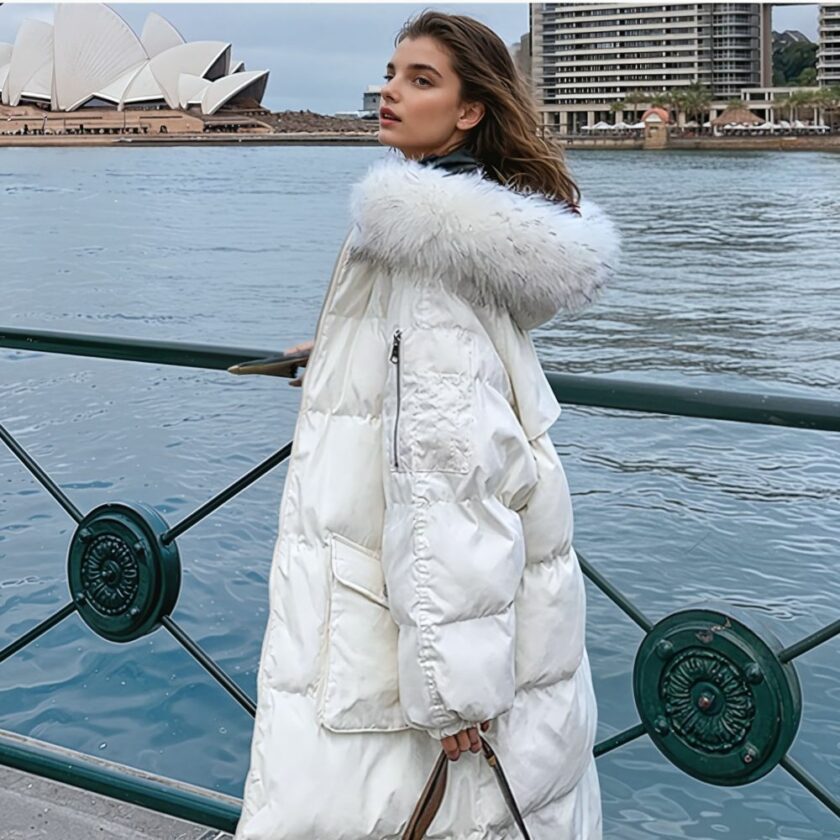 Femme aux cheveux bruns debout à gauche d'une barrière en métal vert. A droite de cette barrière une étendue d'eau, style fleuve et en fond des bâtiment. La femme porte une doudoune longue blanche avec capuche en fourrure blanche. Une lanière de sac dans sa main gauche. Elle est de profil.