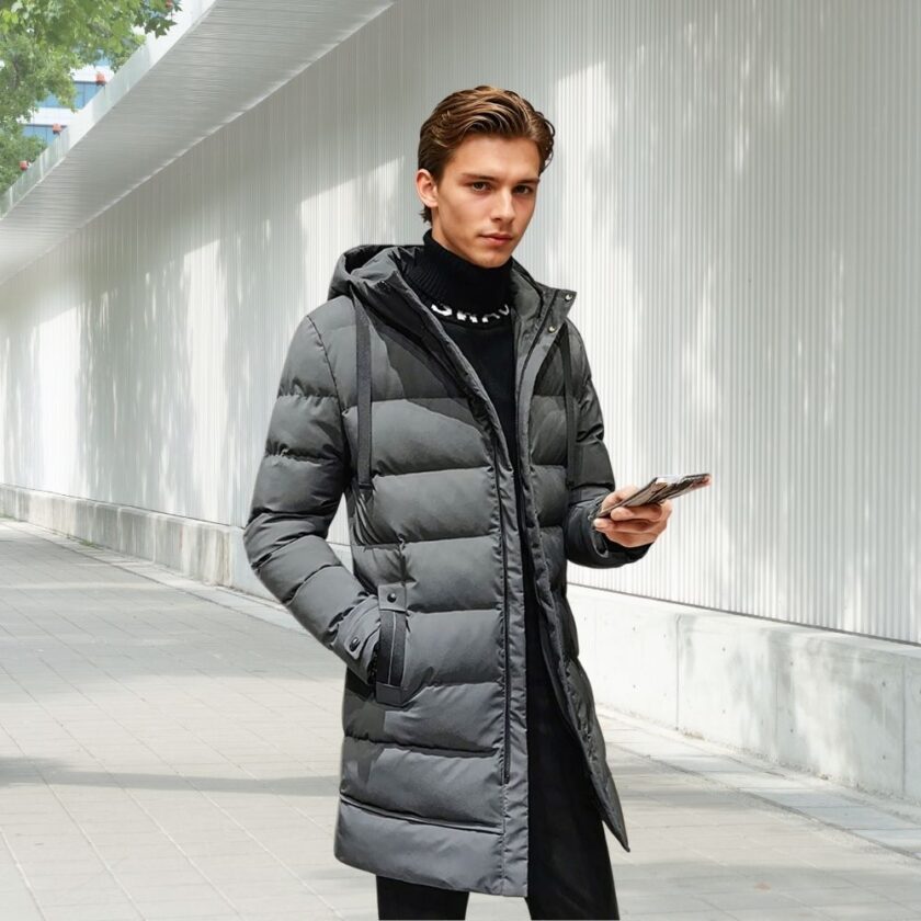 Doudoune en duvet mi-longue pour homme en version grise et portée par un jeune homme aux cheveux brun et qui tient son téléphone dans la main gauche. Il se tient debout dans la rue.