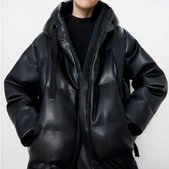 On voit le buste d'une femme blanche qui porte une doudoune oversize en similicuir noir. Elle a les mains dans les poches. Le fond de la photo est gris clair.