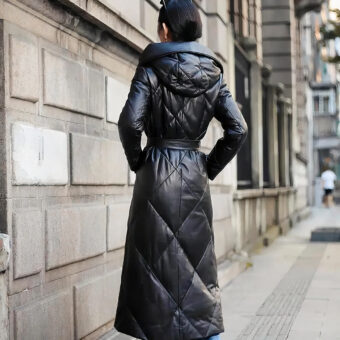 On voit une femme brune de dos, dans la rue. Elle porte un chignon. Elle porte une doudoune longue en similicuir noir. Elle a les mains dans les poches. Elle se trouve près d'un mur très joli, typique de Paris, ou d'une grande ville à la belle architecture.