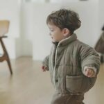Petit garçon qui porte la doudoune bébé doublée polaire épaisse et chaude dans sa version kaki. Ce petit garçon la porte à l'intérieur de sa maison, dans ce qui semble être sa salle à manger.