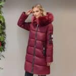 Femme blonde de profil à côté d'un mur marron clair portant une doudoune chaude mi-longue de couleur bordeaux avec de la fourrure assortie sur la capuche. des poches latérales. Elle tient sa main droite à côté de sa tempe. On aperçoit une plante verte à gauche.