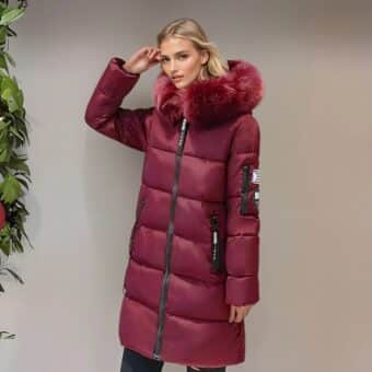 Femme blonde de profil à côté d'un mur marron clair portant une doudoune chaude mi-longue de couleur bordeaux avec de la fourrure assortie sur la capuche. des poches latérales. Elle tient sa main droite à côté de sa tempe. On aperçoit une plante verte à gauche.