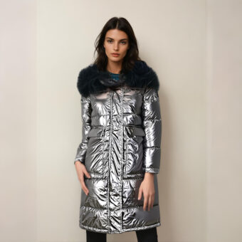 Femme brune dos à un mur beige, portant un pantalon noir et une doudoune brillante et grise mi-longue avec fourrure noire sur la capuche. Deux poches carrées latérales.