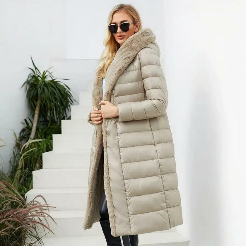 Femme portant une doudoune longue matelassée beige avec col en fourrure beige, fermée par une ceinture. Elle a des cheveux longs détachés et lunettes de soleil avec un sac noir à la main avec des escaliers blancs montants en arrière-plan .