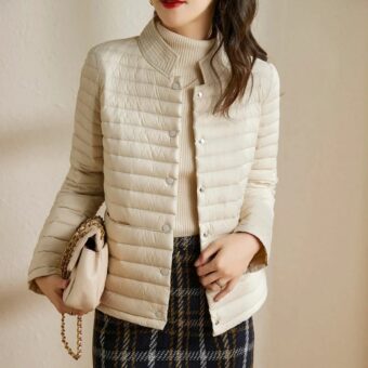 Femme sur fond beige qui porte la doudoune fine à l'allure chic couleur beige au-dessus d'un sous-pull beige et d'une jupe à carreaux avec un petit sac à main beige avec une anse en chaine or posé contre sa hanche et tenu à la main.
