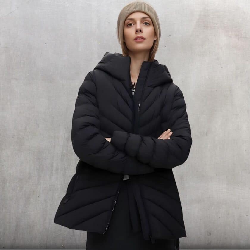 Femme qui porte la doudoune chaude mi-longue matelassée noire les bras croisés avec un bonnet marron sur la tête.